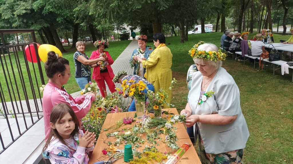 Seniorzy przed budynkiem przygotowują dekoracje z kwiatów na piknik.