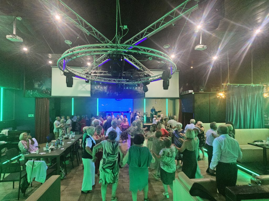 Uczestnicy tańczą w parach i również w grupie. W tle zespół muzyczny.